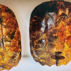 May include: Two wall sconces with a tortoise shell design. The sconces are brown and gold with a textured surface. Each sconce has a small, dark metal candle holder attached. The sconces are designed to be mounted on a wall.