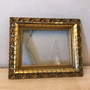 K&ouml;nnte beinhalten: Ein verzierter Bilderrahmen in Gold mit einem dekorativen, geschnitzten Design. Der Rahmen ist aus Holz und hat eine Glasfront.