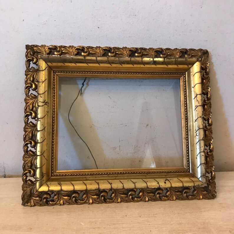K&ouml;nnte beinhalten: Ein verzierter Bilderrahmen in Gold mit einem dekorativen, geschnitzten Design. Der Rahmen ist aus Holz und hat eine Glasfront.