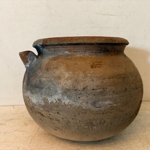 May include: A rustic, round, earthenware pot with a small spout. The pot has a wide opening and a dark brown and gray color scheme. The exterior shows signs of wear, with a textured surface and white patches.