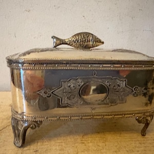 May include: Silver-toned rectangular jewelry box with a fish-shaped handle. The box has ornate detailing, including decorative patterns and curved legs. The lid is slightly domed, and the box appears to be vintage.