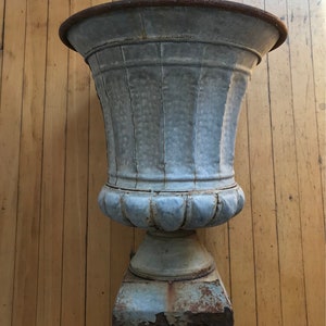 May include: A large, gray metal planter with a fluted design and a pedestal base. The planter has a rustic, weathered look and is made of galvanized metal.