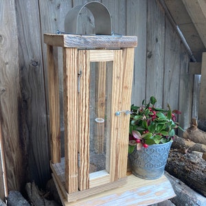 Può includere: Una lanterna in legno rustico con un top in metallo e pannelli in vetro. La lanterna è appoggiata su una superficie in legno con una piccola pianta in vaso davanti a essa.