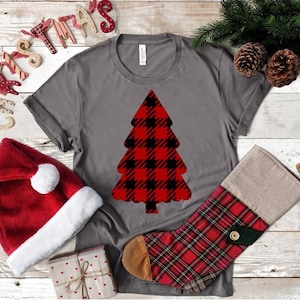 May include: Gray t-shirt with a red and black plaid Christmas tree design. The shirt is laying on a white wooden surface with a red Santa hat and a red and black plaid stocking.