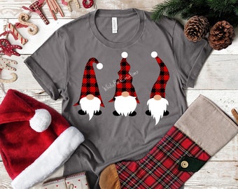 Könnte beinhalten: Drei Gnome mit roten und schwarzen Buffalo-Plaid-Hüten stehen in einer Reihe auf einem grauen T-Shirt. Die Gnome haben weiße Bärte und weiße Gesichter. Der Text "White Buck Creations" ist auf das Shirt gedruckt.