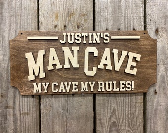 May include: A wooden sign with the text "Justin's Man Cave My Cave My Rules!" in white lettering. The sign has a brown woodgrain finish and a distressed look.