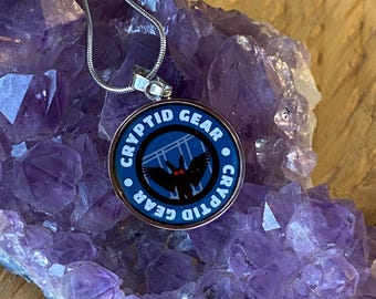 May include: A silver chain necklace with a round pendant featuring a blue background with a black bat with red eyes. The pendant has the text "Cryptid Gear" written around the edge.