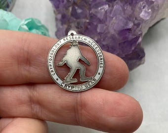May include: A silver pendant featuring a Bigfoot design. The circular pendant has the text "CRYPTOZOOLOGY RESEARCH ACCESSORIES" and "CRYPTID GEAR" around the edge, with a Bigfoot silhouette in the middle.