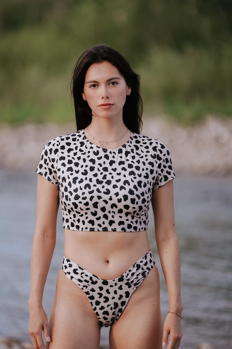May include: A two-piece swimsuit featuring a short-sleeved crop top and high-cut bikini bottoms. Both pieces are made of a light beige fabric covered in a black animal print pattern. The top has a round neckline and the bottoms have a high-leg cut.