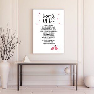 May include: A white framed print with a black and pink design. The print says "Heiratsantrag" and has a poem in German. The print is decorated with pink hearts and two pink birds.