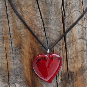 Op de afbeelding: Een rode glazen hartvormige hanger aan een zwarte koordsnoer. De ketting hangt aan een stuk verweerd hout.