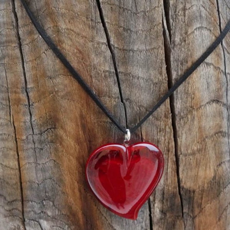 Op de afbeelding: Een rode glazen hartvormige hanger aan een zwarte koordsnoer. De ketting hangt aan een stuk verweerd hout.