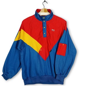 Könnte beinhalten: Ein Vintage-Pullover mit Farbblock-Design in Rot, Gelb und Blau. Das langärmlige Oberteil hat einen hohen Kragen, einen Knopfverschluss und eine kleine Tasche. Der Pullover hat eine Retro-Ästhetik.