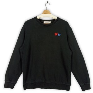 Può includere: Felpa nera girocollo con logo ricamato a forma di cuore rosso e blu. La felpa ha maniche lunghe e orlo e polsini a coste. Il marchio "C.D.G. PLAY" è stampato sul colletto.
