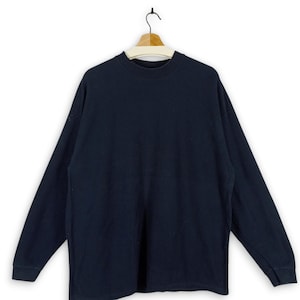 Puede incluir: Una camisa de manga larga azul oscuro colgada de una percha de madera. La camisa tiene cuello redondo y está hecha de una tela suave y cómoda.