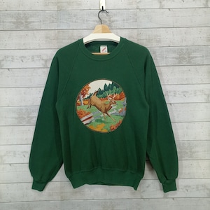 Op de afbeelding: Een donkergroen sweatshirt met een ronde afbeelding van een hert dat door een boslandschap rent. Het hert is bruin met geweien en wordt omringd door bomen en bladeren.