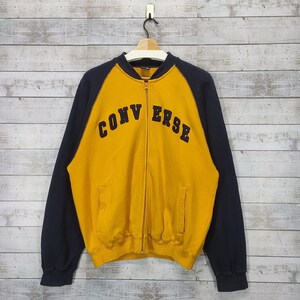 Op de afbeelding: Een geel en zwart ritsjack met het woord "CONVERSE" in zwarte letters op de voorkant.