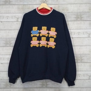 May include: A navy blue sweatshirt with a crew neck and a red and white striped collar. The sweatshirt features a design of eight brown teddy bears wearing American flag shirts.