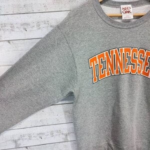 May include: Gray sweatshirt with a large orange "Tennessee" graphic on the front. The sweatshirt has a crew neck and long sleeves. The tag on the sweatshirt says "Red Oak".