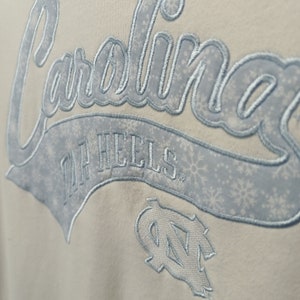 May include: A white sweatshirt with a blue embroidered design that says "Carolina Tar Heels" with a snowflake pattern. The design also includes a blue embroidered "UNC" logo.