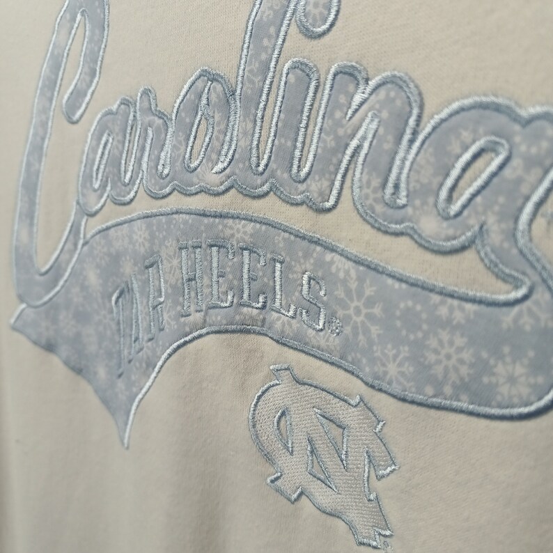 May include: A white sweatshirt with a blue embroidered design that says "Carolina Tar Heels" with a snowflake pattern. The design also includes a blue embroidered "UNC" logo.