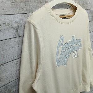 May include: A cream colored sweatshirt with a blue embroidered graphic that says "Carolina Tar Heels" with a blue and white logo.