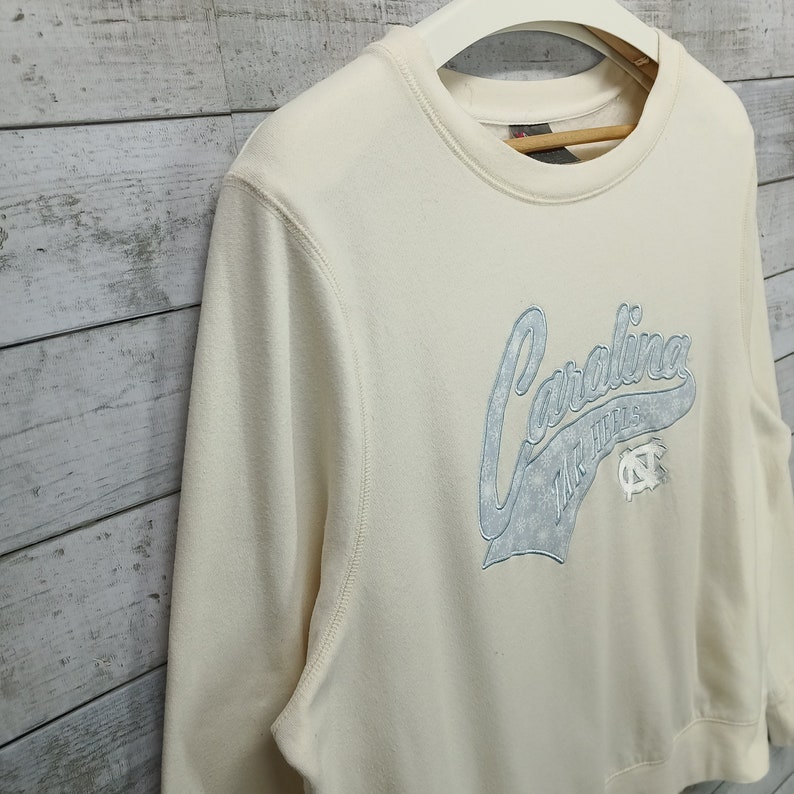 May include: A cream colored sweatshirt with a blue embroidered graphic that says "Carolina Tar Heels" with a blue and white logo.