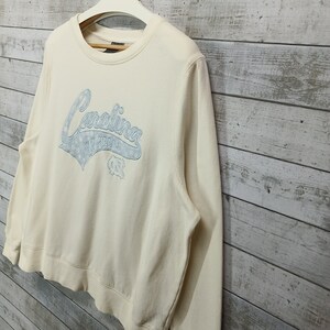 May include: A cream colored sweatshirt with a blue and white Carolina Tar Heels logo on the front.
