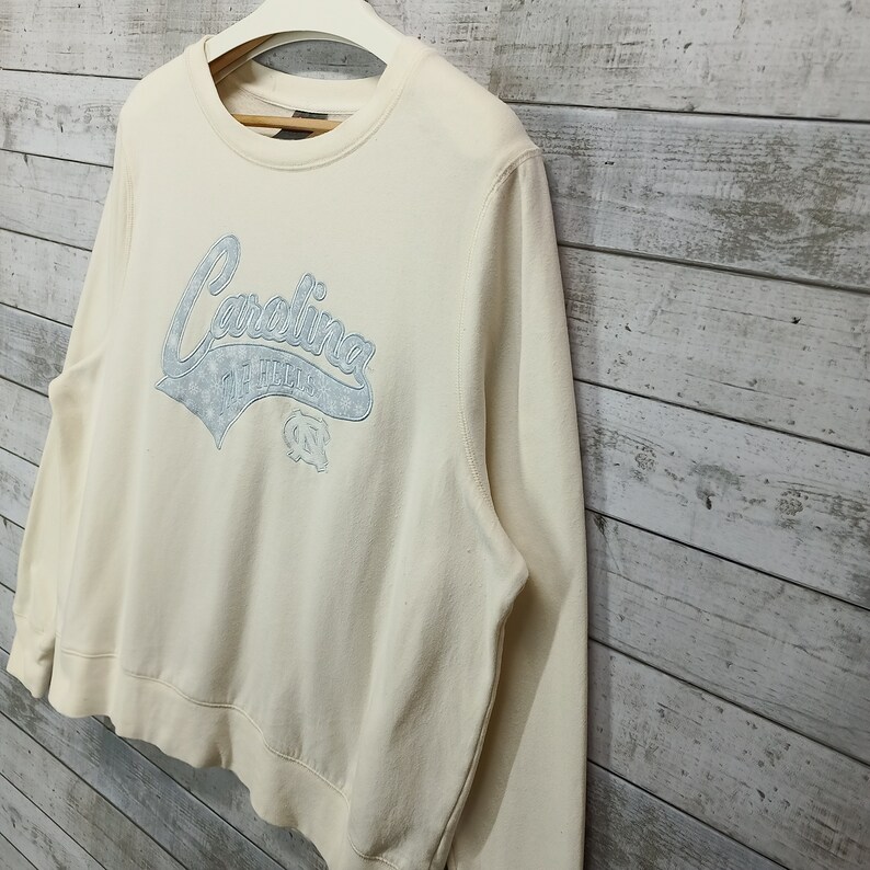 May include: A cream colored sweatshirt with a blue and white Carolina Tar Heels logo on the front.