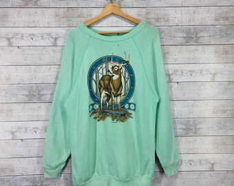 May include: A light green sweatshirt with a graphic of a deer in a forest scene. The deer is brown with antlers and is standing in a circle with a blue border. The graphic is surrounded by a brown ribbon with the words "The Outdoorsman" printed on it.