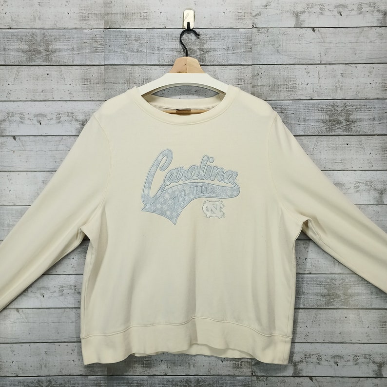 May include: A cream colored long-sleeved sweatshirt with a blue embroidered graphic that says "Carolina" with a star design and a blue "NC" logo.
