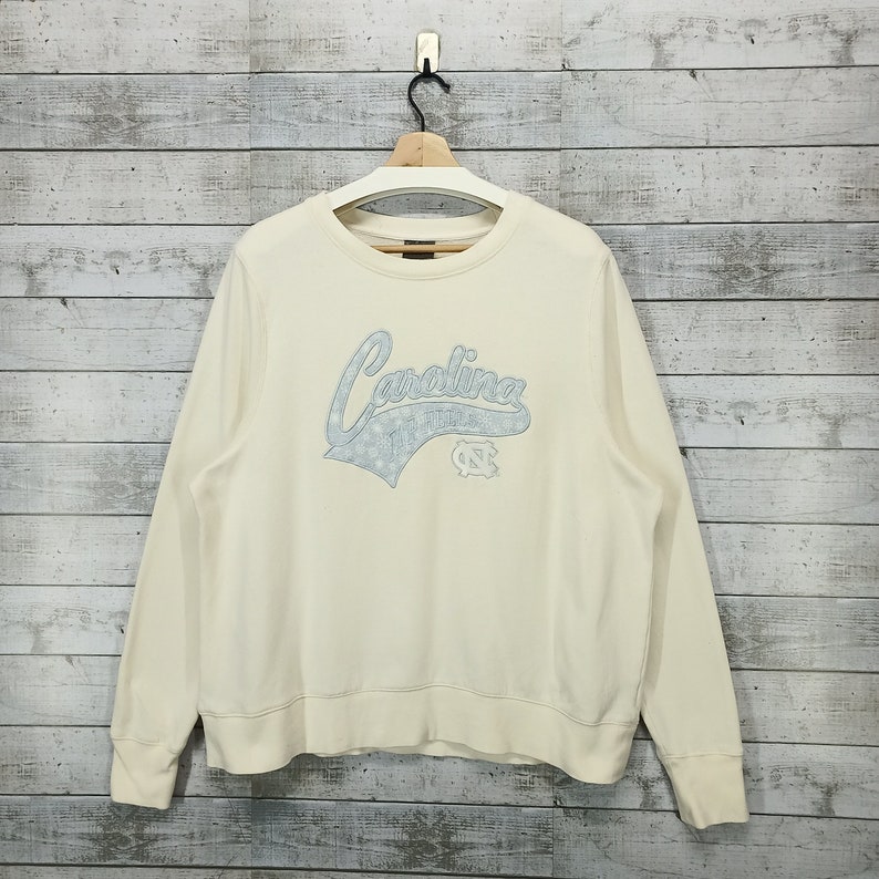 May include: A cream-colored sweatshirt with a blue and white Carolina Tar Heels logo on the front.