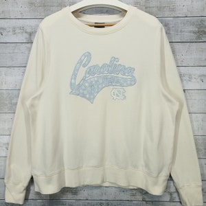 May include: A cream colored sweatshirt with a blue and white Carolina Tar Heels logo. The logo features the word "Carolina" in a script font with snowflakes and the Tar Heels logo.