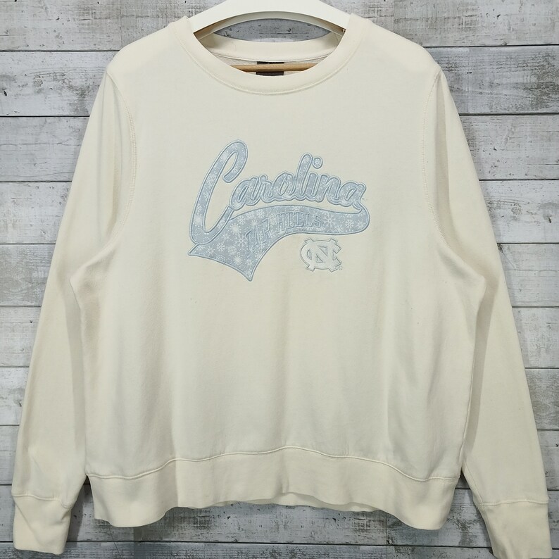 May include: A cream colored sweatshirt with a blue and white Carolina Tar Heels logo. The logo features the word "Carolina" in a script font with snowflakes and the Tar Heels logo.