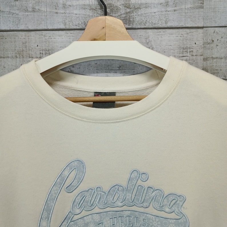May include: A cream colored sweatshirt with a blue embroidered logo that says "Carolina" with snowflakes around the text.