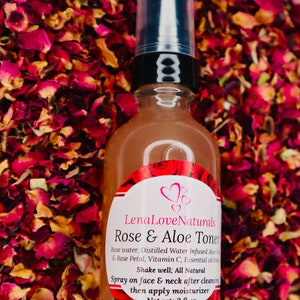 May include: A clear glass bottle with a black pump top and a white label. The label says "Lena Love Naturals Rose & Aloe Toner" and lists the ingredients as "Rose water, Distilled Water Infused Aloe Vera & Rose Petal, Vitamin C, Essential oil blend". The label also says "Shake well; All Natural" and "Spray on face & neck after cleansing then apply moisturizer". The bottle is sitting on a bed of red rose petals.