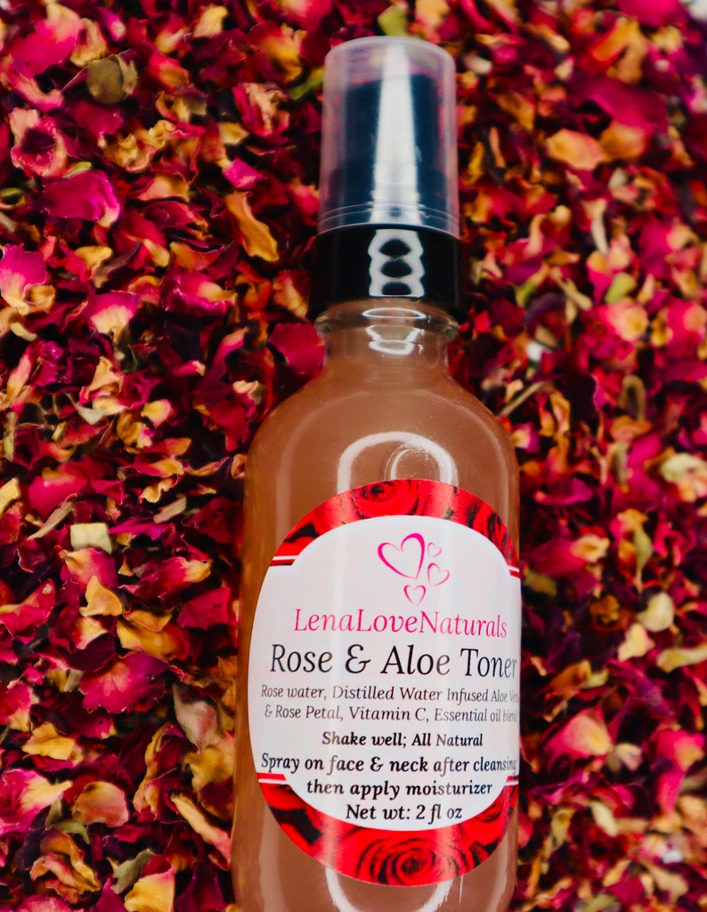 May include: A clear glass bottle with a black pump top and a white label. The label says "Lena Love Naturals Rose & Aloe Toner" and lists the ingredients as "Rose water, Distilled Water Infused Aloe Vera & Rose Petal, Vitamin C, Essential oil blend". The label also says "Shake well; All Natural" and "Spray on face & neck after cleansing then apply moisturizer". The bottle is sitting on a bed of red rose petals.