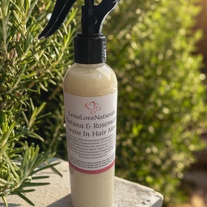 May include: A clear bottle of LenaLoveNaturals Batana & Rosemary Leave In Hair Mist. The bottle has a black spray nozzle and a white label with text and a heart design. The background is blurred green foliage.