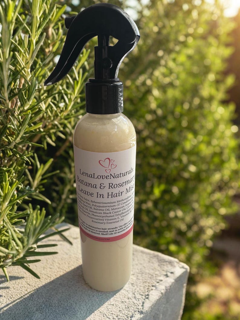 May include: A clear bottle of LenaLoveNaturals Batana & Rosemary Leave In Hair Mist. The bottle has a black spray nozzle and a white label with text and a heart design. The background is blurred green foliage.