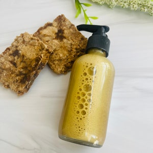 May include: A clear glass bottle with a black pump dispenser filled with a light brown liquid. The liquid has small bubbles throughout. Two pieces of brown soap are in the background.