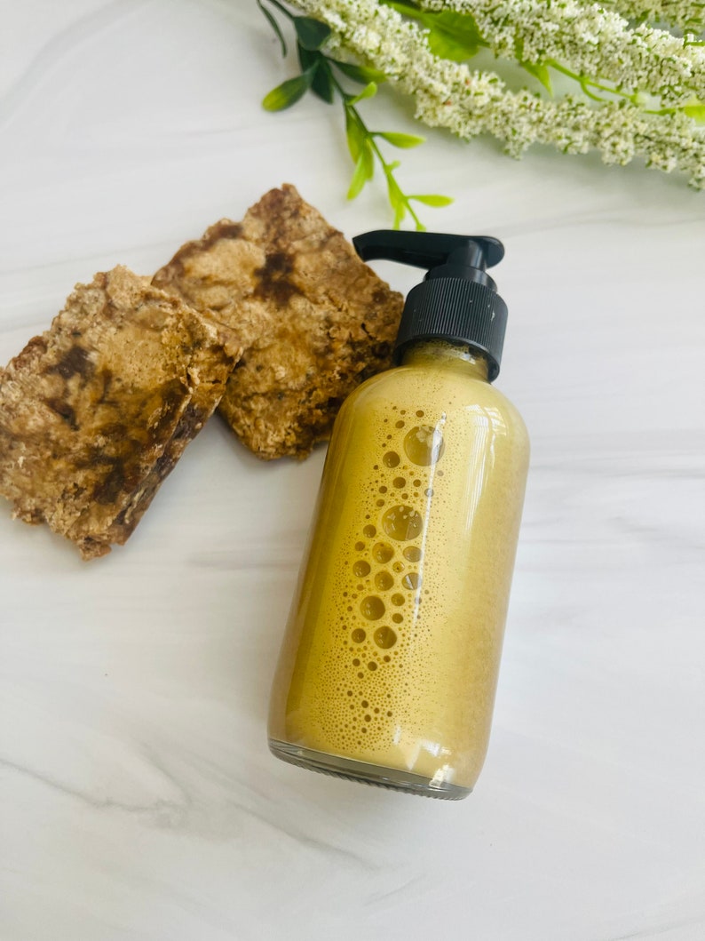 May include: A clear glass bottle with a black pump dispenser filled with a light brown liquid. The liquid has small bubbles throughout. Two pieces of brown soap are in the background.