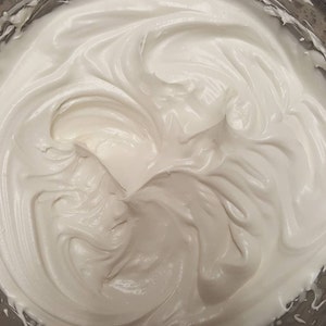 May include: A close-up of a white meringue mixture in a silver mixing bowl. The meringue is smooth and fluffy, with swirls and peaks.