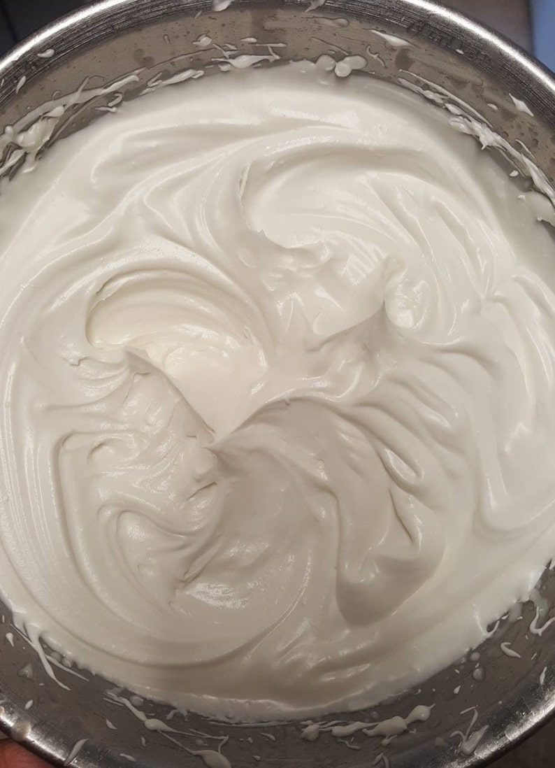 May include: A close-up of a white meringue mixture in a silver mixing bowl. The meringue is smooth and fluffy, with swirls and peaks.