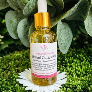 May include: A clear glass bottle of LenaLoveNaturals Herbal Cuticle Oil with a gold cap and white dropper. The label lists ingredients like Sweet Almond Oil and French Lavender Oil. The bottle sits on a white flower against a green background.