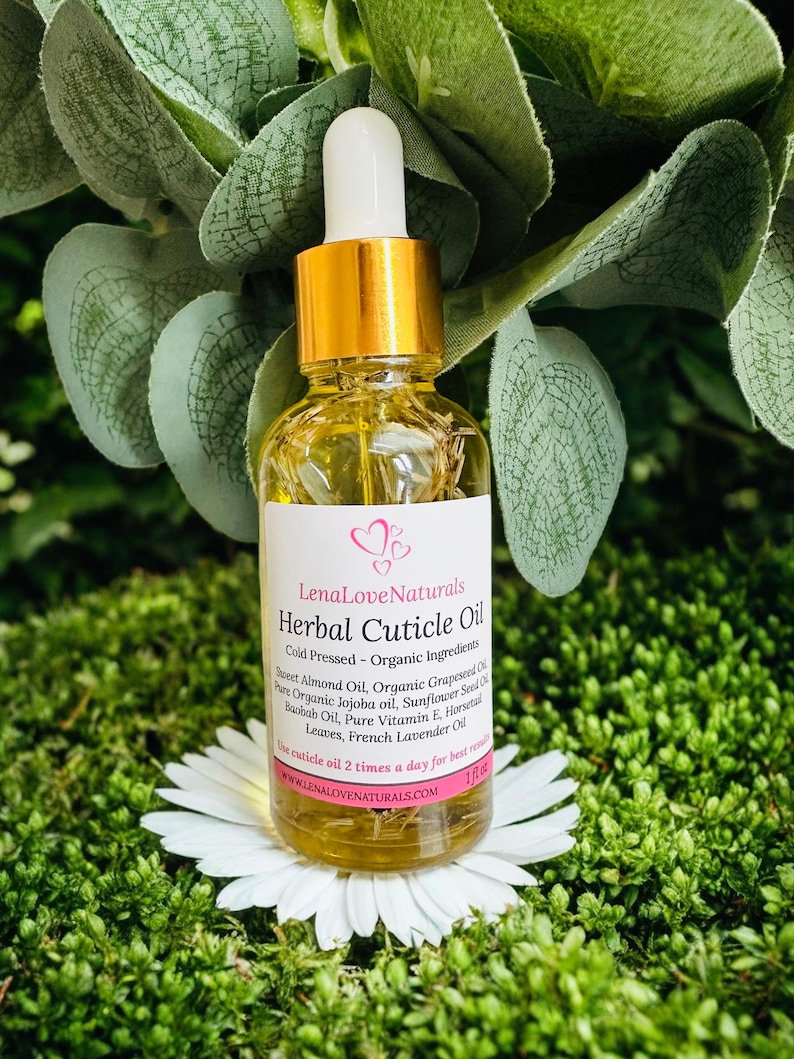 May include: A clear glass bottle of LenaLoveNaturals Herbal Cuticle Oil with a gold cap and white dropper. The label lists ingredients like Sweet Almond Oil and French Lavender Oil. The bottle sits on a white flower against a green background.