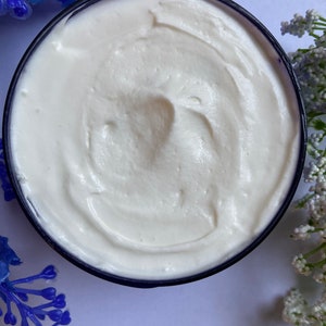 May include: A close-up of a white, creamy body butter in a blue jar. The butter has a smooth, whipped texture.