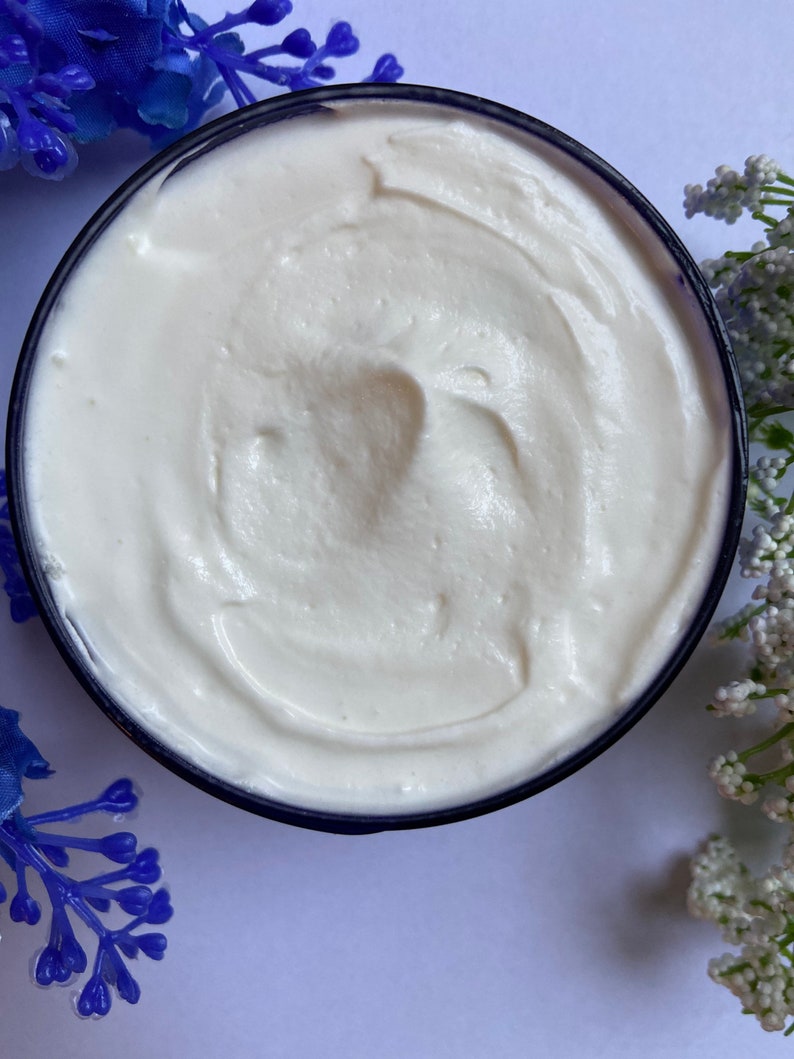 May include: A close-up of a white, creamy body butter in a blue jar. The butter has a smooth, whipped texture.