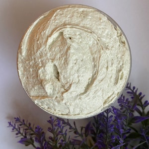 May include: A close-up of a white, creamy body butter in a clear container. The butter has a smooth, whipped texture. Lavender flowers are in the background.