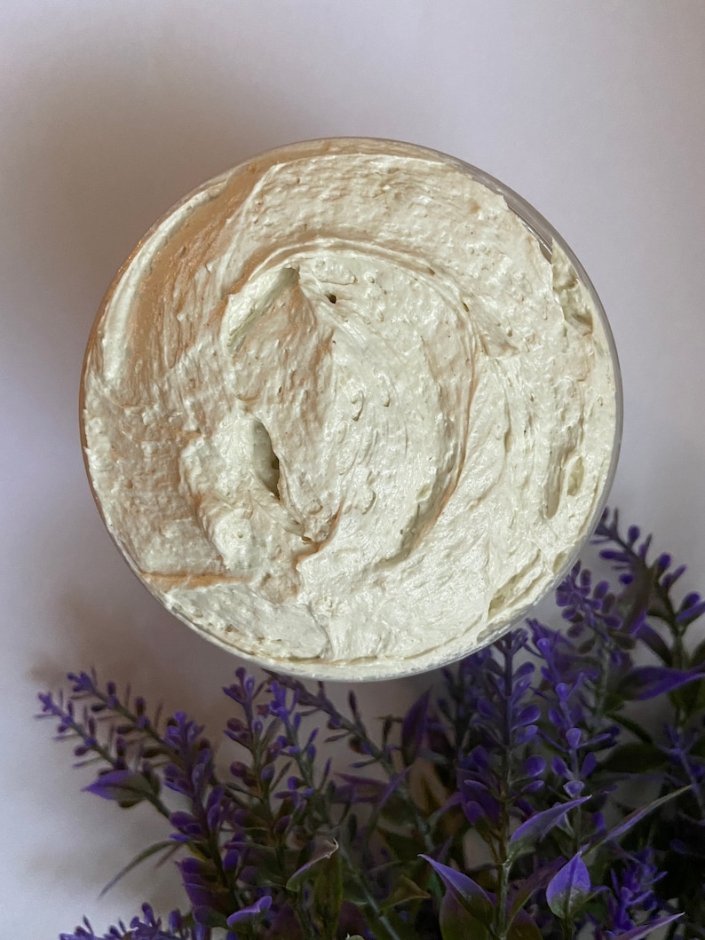 May include: A close-up of a white, creamy body butter in a clear container. The butter has a smooth, whipped texture. Lavender flowers are in the background.