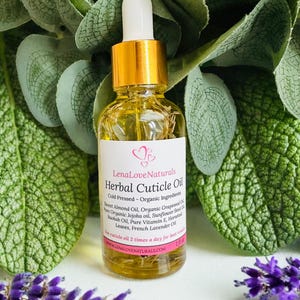 Op de afbeelding: Een heldere glazen fles Lena Love Naturals Herbal Cuticle Oil met een gouden dop en witte druppelaar. Het etiket vermeldt ingrediënten zoals zoete amandelolie en Franse lavendel. De fles is omgeven door groene bladeren en paarse bloemen.